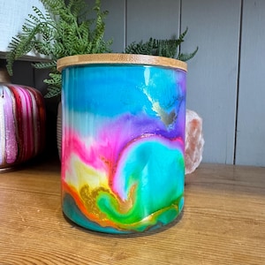 Puede incluir: Un recipiente de almacenamiento cilíndrico con un vibrante patrón abstracto de remolinos en colores turquesa brillante, rosa intenso, morado, verde y naranja con detalles dorados. El recipiente tiene una tapa de bambú natural claro.
