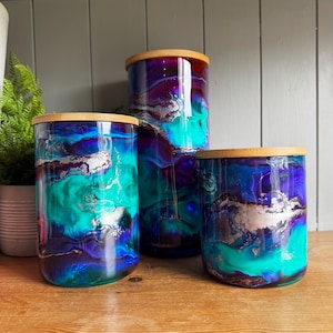 May include: Three cylindrical glass canisters of varying heights, each featuring a vibrant marbled pattern of deep blue, bright turquoise, and metallic silver swirls. Each canister is topped with a circular, light-coloured bamboo lid.