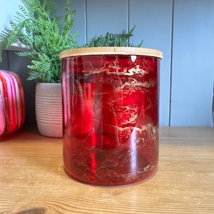 May include: A red glass jar with gold marbling and a wooden lid. The jar is cylindrical and sits on a wooden surface. A green plant in a white pot and a pink and gold object are in the background.