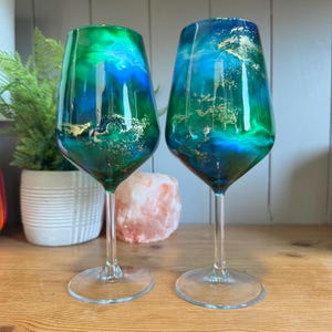 Op de afbeelding: Twee wijnglazen met een opvallend abstract ontwerp. De glazen tonen een mix van groene en blauwe tinten, geaccentueerd met bladgoud. De heldere stelen en bases vormen een delicaat contrast met de kleurrijke kelken.