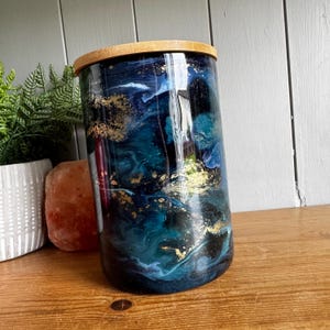 May include: A cylindrical glass jar featuring a glossy finish with a swirling marble pattern in shades of deep navy blue, teal, and black, accented with metallic gold flakes. The container is topped with a circular, light-coloured bamboo lid.