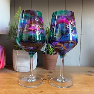 May include: A pair of wine glasses with a colourful, abstract design. The glasses have a mix of teal, purple, and gold tones, creating a galaxy-like effect. The clear stems and bases contrast with the vibrant bowls.