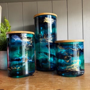 May include: Three cylindrical glass storage canisters of varying heights, each featuring a deep blue and teal marbled pattern with gold leaf accents and a light-coloured wooden lid. The glass surfaces display swirling colours and metallic details.