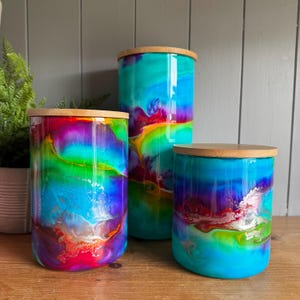 May include: Three cylindrical storage canisters of varying heights, each featuring a vibrant, swirling alcohol ink pattern in shades of turquoise, magenta, purple, and yellow, topped with a circular, light-coloured bamboo lid.