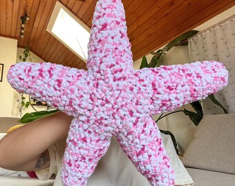 Puede incluir: Un cojín de felpa con forma de estrella de mar de cinco puntas, elaborado con hilo grueso y suave. La textura es densa y con bucles, presentando un patrón de colores variados en blanco, rosa claro y rosa oscuro en toda la superficie.
