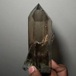 May include: A large, translucent smoky quartz crystal point with a sharp, faceted termination. The crystal displays a deep brown, semi-transparent colour with visible internal inclusions and natural geometric facets along its vertical structure.
