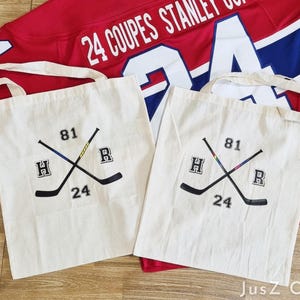 Puede incluir: Dos bolsas de mano color crema con asas blancas, cada una con un diseño de palo de hockey negro con los números 81 y 24. Una camiseta de hockey roja con texto blanco en el fondo.