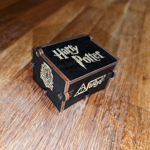 May include: A black wooden box featuring the words "Harry Potter" and "Always" engraved on it. The box has a decorative crest and a lightning bolt design. The box is on a wooden surface.