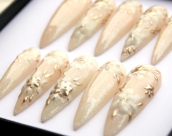 Könnte beinhalten: Ein Set aus zehn langen, mandelförmigen Kunstnägeln. Jeder Nagel ist ein blasses, schimmerndes Beige mit goldenen Blumen- und Blattakzenten. Die Nägel sind in einer schwarzen Box angeordnet, die das aufwendige Design zeigt.