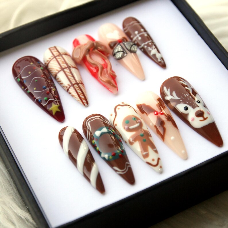 May include: A set of ten almond-shaped artificial nails with festive holiday designs. The nails feature various patterns, including a Christmas tree, a gingerbread man, and a reindeer, in shades of brown, red, white, and beige. The nails are displayed in a black box.