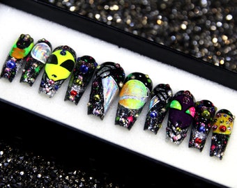 May include: A set of ten black press-on nails with colorful alien and planet designs. The nails are decorated with rhinestones and glitter.