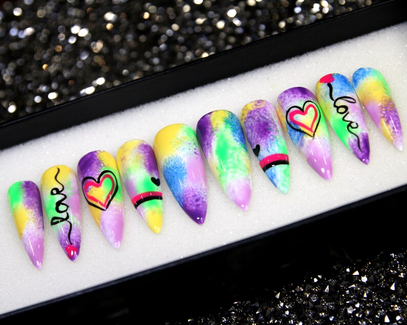 May include: Set of ten colorful, long, pointed false nails with a tie-dye design. Each nail has a different design, including hearts, swirls, and the word "love".