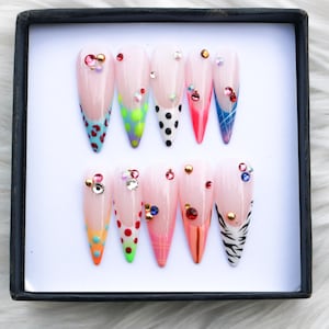 May include: A set of ten almond-shaped press-on nails featuring a nude base with diverse colorful tips including leopard print, polka dots, plaid, and zebra stripes. Each nail is embellished with various small metallic and crystal rhinestones.