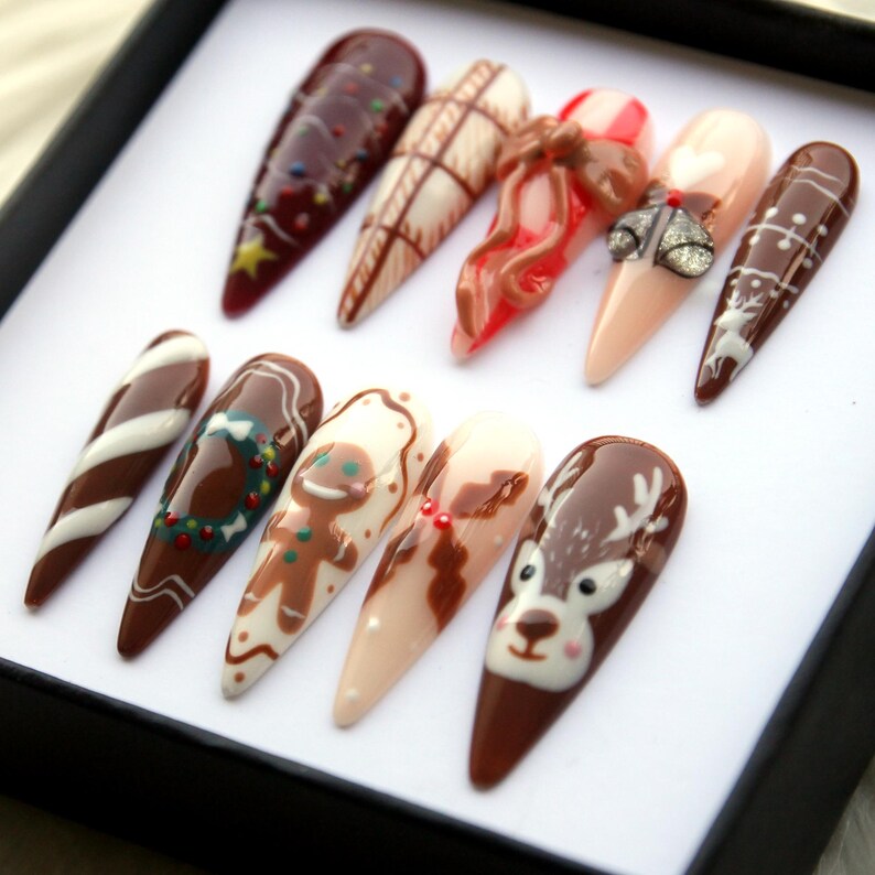 May include: A set of ten long, almond-shaped artificial nails with various Christmas-themed designs. The nails feature colors like brown, white, red, and beige, with designs including a gingerbread man, candy cane stripes, and a reindeer.
