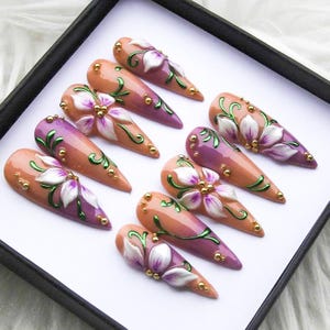 May include: A set of ten almond-shaped artificial nails. Each nail features a gradient design in shades of peach and purple, adorned with white and purple floral accents, green vines, and gold embellishments.