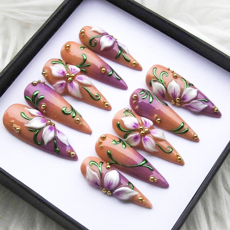 May include: A set of ten almond-shaped artificial nails. Each nail features a gradient design in shades of peach and purple, adorned with white and purple floral accents, green vines, and gold embellishments.