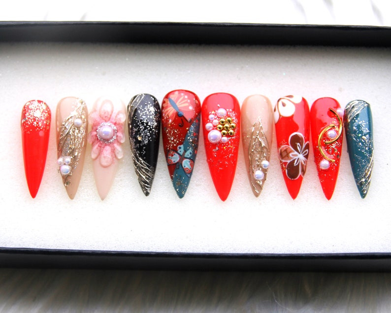 May include: Set of ten false nails with various designs. The nails are red, black, gold, white, and blue with floral and butterfly patterns. Some nails have glitter and pearls.
