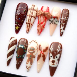 May include: A set of ten long, almond-shaped artificial nails with festive Christmas designs. The nails feature various designs, including a Christmas tree, candy cane, gingerbread man, and reindeer, in shades of brown, red, white, and beige.