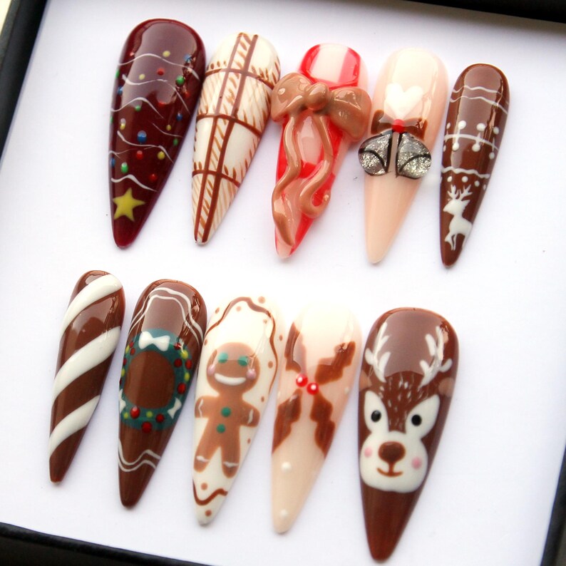 May include: A set of ten long, almond-shaped artificial nails with festive Christmas designs. The nails feature various designs, including a Christmas tree, candy cane, gingerbread man, and reindeer, in shades of brown, red, white, and beige.