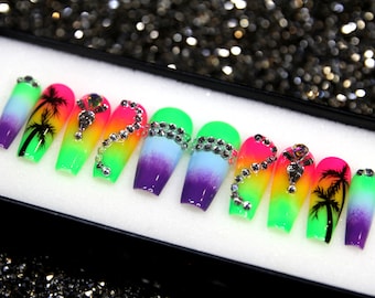 May include: A set of ten colorful, ombre press-on nails with palm tree designs and rhinestones. The nails are in a variety of colors, including green, yellow, pink, blue, and purple. The nails are arranged in a box.