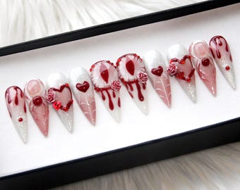 May include: A set of ten long, pointed artificial nails with a spooky theme. Each nail features a unique design, including red dripping blood, hearts, and decorative embellishments. The nails are displayed in a black box against a white, fluffy background.