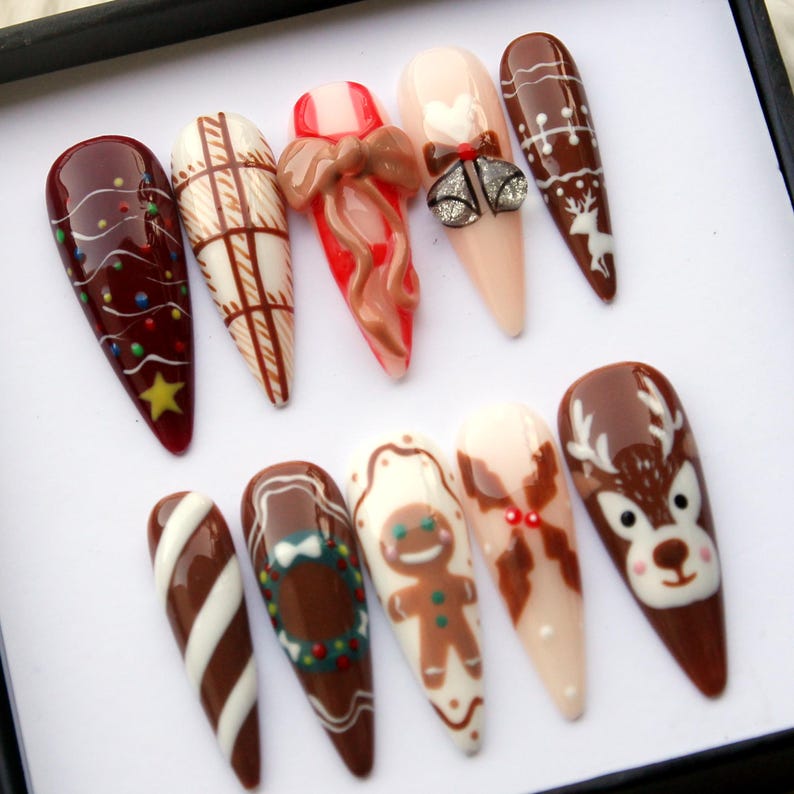 May include: A set of ten long, almond-shaped artificial nails with various Christmas-themed designs. The designs include a Christmas tree, plaid pattern, bow, bells, reindeer, candy cane stripes, gingerbread man, and a deer face. Colors include brown, white, red, and gold.
