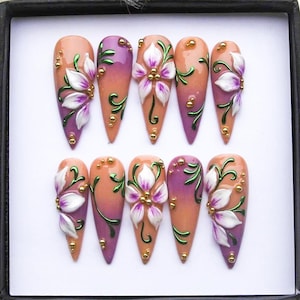 May include: A set of ten long, almond-shaped artificial nails. Each nail features a gradient design, transitioning from peach to purple. They are adorned with white and purple floral accents, green vines, and gold embellishments.