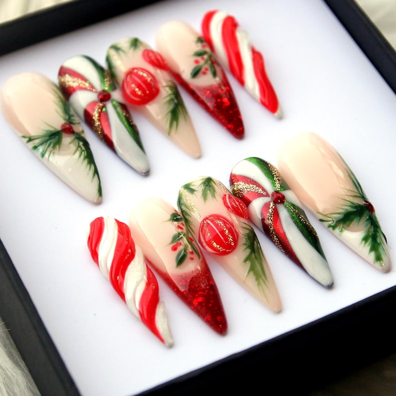 May include: A set of ten almond-shaped artificial nails with festive Christmas designs. The nails feature candy cane stripes, red glitter tips, and hand-painted holly and berry accents. Some nails have a 3D candy embellishment.