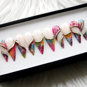 Puede incluir: Un conjunto de uñas postizas en forma de almendra. Cada uña presenta una base blanca con un diseño marmolado colorido en tonos rosa, azul, amarillo y rojo. Un acento dorado curvado adorna cada uña, agregando un toque de elegancia.