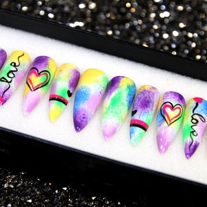 May include: A set of ten colorful, long, stiletto-shaped false nails with a tie-dye design. Each nail has a different design featuring hearts and the word "love" in black and pink lettering.
