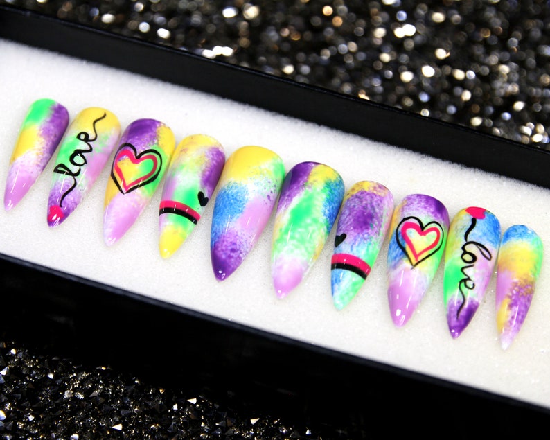 May include: A set of ten colorful, long, stiletto-shaped false nails with a tie-dye design. Each nail has a different design featuring hearts and the word "love" in black and pink lettering.