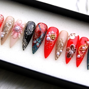 May include: Set of ten false nails with a stiletto shape. The nails are decorated with red, black, blue, and gold glitter, white and gold flowers, and pearls.
