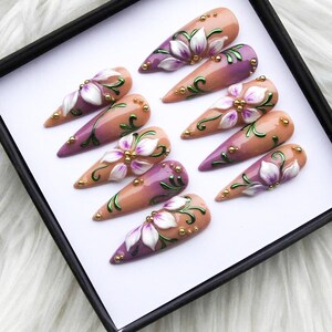 May include: A set of ten long, almond-shaped artificial nails. Each nail features a gradient design, transitioning from a nude base to a purple tip. White floral designs with green vines and gold accents adorn each nail, creating a delicate, artistic look.