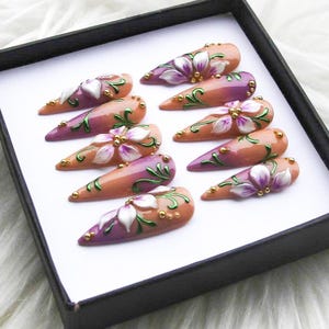 May include: A set of ten almond-shaped false nails. Each nail is painted with a gradient of peach and purple, adorned with white and purple floral designs, green vines, and gold accents. The nails are displayed in a black box.