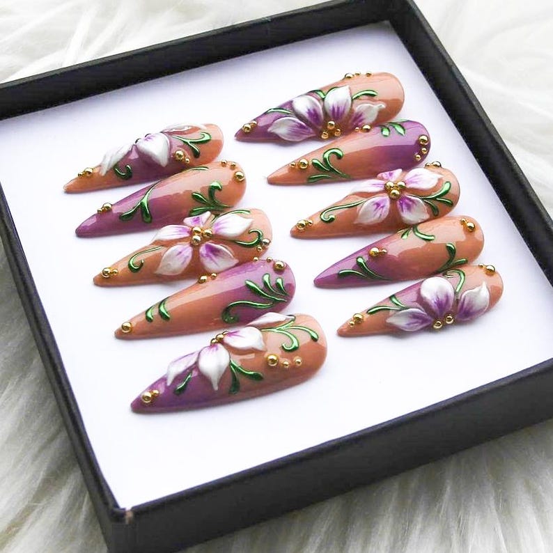 May include: A set of ten almond-shaped false nails. Each nail is painted with a gradient of peach and purple, adorned with white and purple floral designs, green vines, and gold accents. The nails are displayed in a black box.