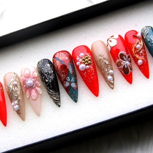 May include: A set of ten false nails with a variety of designs. The nails are red, pink, black, and teal with gold accents and pearl embellishments. The nails are arranged in a row in a white box.