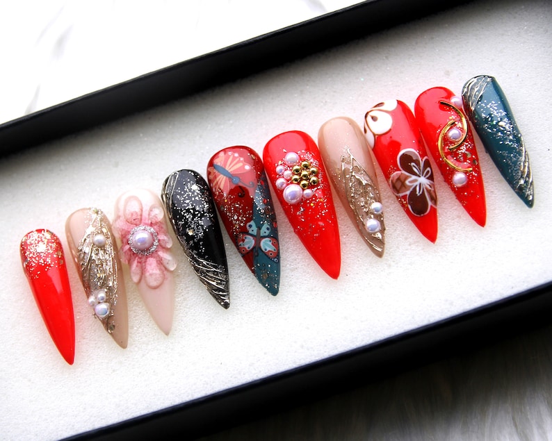 May include: A set of ten false nails with a variety of designs. The nails are red, pink, black, and teal with gold accents and pearl embellishments. The nails are arranged in a row in a white box.