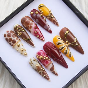 Puede incluir: Una colección de uñas postizas largas en forma de almendra. Los diseños incluyen lunares, remolinos y colores lisos en tonos marrón, rojo y amarillo. Adornos con cuentas doradas y detalles decorativos.