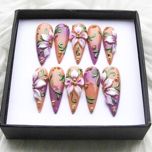 May include: A set of ten artificial nails, each with a unique design. The nails feature a gradient of peach and purple hues, adorned with white and purple floral embellishments, green vines, and gold accents. They are presented in a black box.