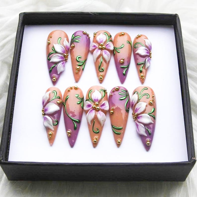 May include: A set of ten artificial nails, each with a unique design. The nails feature a gradient of peach and purple hues, adorned with white and purple floral embellishments, green vines, and gold accents. They are presented in a black box.