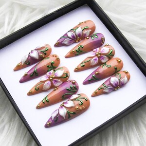 May include: A set of ten almond-shaped artificial nails. Each nail features a gradient of peach and purple, adorned with a white and purple floral design, green vines, and gold accents. The nails are displayed in a black box.