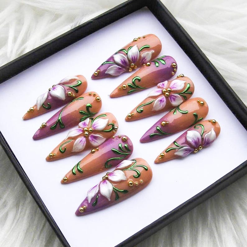 May include: A set of ten almond-shaped artificial nails. Each nail features a gradient of peach and purple, adorned with a white and purple floral design, green vines, and gold accents. The nails are displayed in a black box.