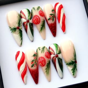 May include: A set of ten artificial nails with a Christmas theme. Designs include red and white candy cane stripes, green holly leaves, red berries, and glitter. Some nails feature a swirl design with red, white, and green stripes, accented with gold glitter.