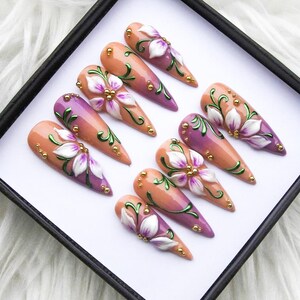 May include: A set of ten long, almond-shaped artificial nails. Each nail features a gradient design, transitioning from peach to purple, adorned with white floral accents, green vines, and gold embellishments.
