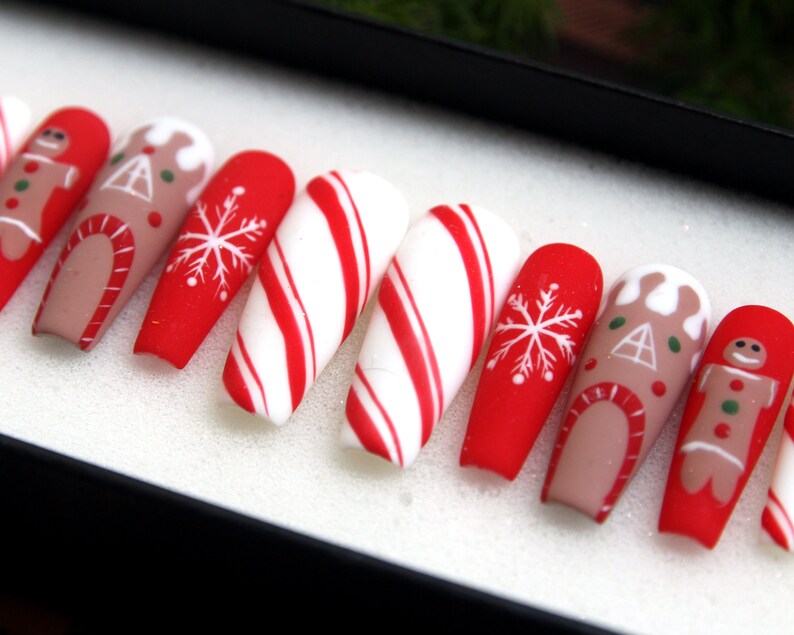 Puede incluir: Conjunto de diez u&ntilde;as postizas con tem&aacute;tica navide&ntilde;a. Las u&ntilde;as est&aacute;n decoradas con hombres de jengibre, casas de jengibre, bastones de caramelo y copos de nieve. Las u&ntilde;as son rojas, blancas y marrones.