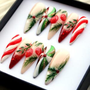 May include: A set of ten long, almond-shaped artificial nails. The nails feature festive designs, including candy cane stripes, green holly leaves, red berries, and glitter accents. The color palette includes red, white, green, and nude tones, perfect for the holiday season.