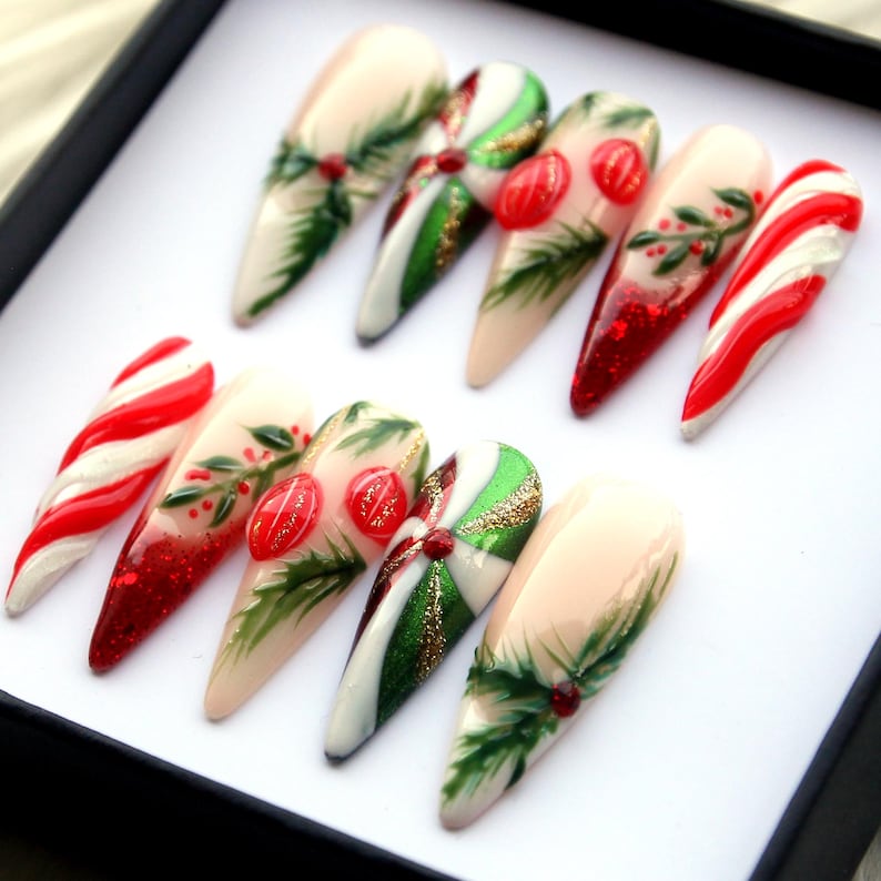 May include: A set of ten long, almond-shaped artificial nails. The nails feature festive designs, including candy cane stripes, green holly leaves, red berries, and glitter accents. The color palette includes red, white, green, and nude tones, perfect for the holiday season.