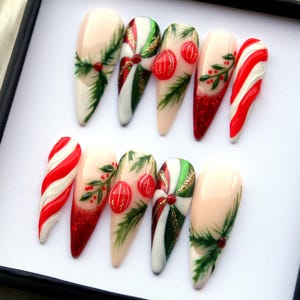 May include: A set of ten long, almond-shaped artificial nails. The nails feature Christmas-themed designs, including candy cane stripes, red glitter, green sprigs, and red ornaments. The base color is a nude pink.