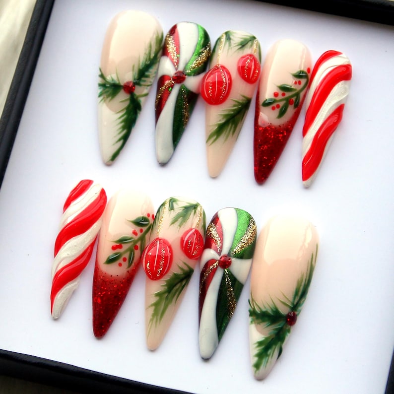 May include: A set of ten long, almond-shaped artificial nails. The nails feature Christmas-themed designs, including candy cane stripes, red glitter, green sprigs, and red ornaments. The base color is a nude pink.