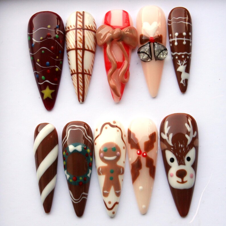 May include: A collection of ten long, almond-shaped artificial nails with various Christmas-themed designs. The designs include a gingerbread man, reindeer, bows, and candy cane stripes, in shades of brown, red, white, and beige.